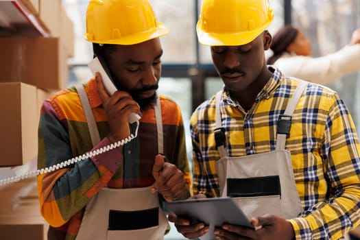 African american storehouse employees coordinating delivery schedule with logistics manager on landline telephone. Warehouse operator talking on phone and looking at digital tablet screen