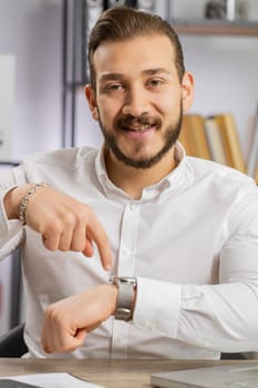 It is your time. Young boss business man showing time on wrist watch clock, approve, pointing finger, time to start work, deadline. Freelancer guy at office workplace looking at camera. Vertical shot