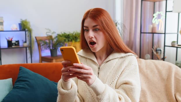 Happy young woman use mobile smartphone typing browsing say Wow yes found out great big win good news celebrate lottery jackpot doing winner gesture. Redhead girl at home in room sitting on sofa