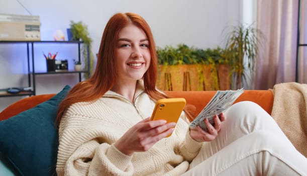 Planning family budget. Smiling redhead woman counting money cash, use smartphone calculate domestic bills at home room. Joyful girl satisfied of income and saves money for planned vacation, gifts