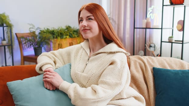 Redhead happy calm young woman at home couch smiling friendly, glad expression looking away dreaming resting, relaxation feel satisfied concept good news, celebrate win. Girl on living room sofa