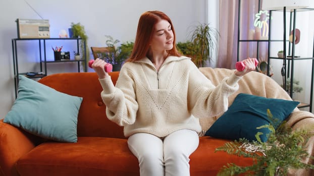 Woman doing dumbbells weight lifting biceps excersices at home. Workout, burning fat calories, training strength, practicing domestic fitness aerobics. Sport motivation. Redhead girl sitting on sofa