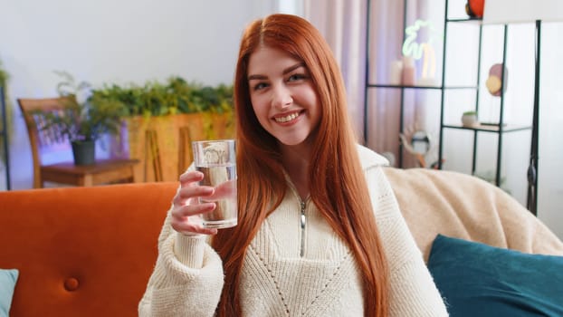 Thirsty redhead woman holding glass of natural aqua make sips drinking still water preventing dehydration sits at home living room. Girl with good life habits, healthy slimming, weight loss concept