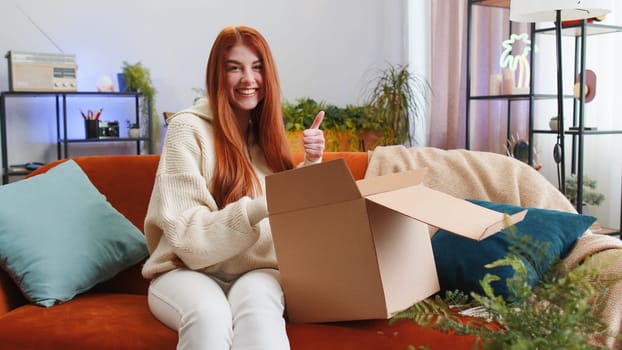 Happy young woman unpacking delivery parcel sitting at home apartment on sofa. Smiling satisfied girl shopper online shop customer opening cardboard box receiving purchase gift by fast postal shipping