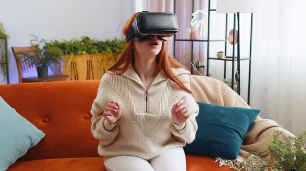 Redhead woman using virtual reality futuristic technology VR app headset helmet to play simulation 3D 360 video game, watching film movie at modern home apartment. Girl in goggles sitting on couch