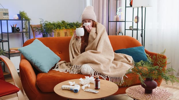 Young ill woman suffering from cold or allergy sitting on sofa in living room at home drinking hot tea. Sick redhead girl wear hat wrapped in plaid blows sneezes wipes snot into napkin. Quarantine