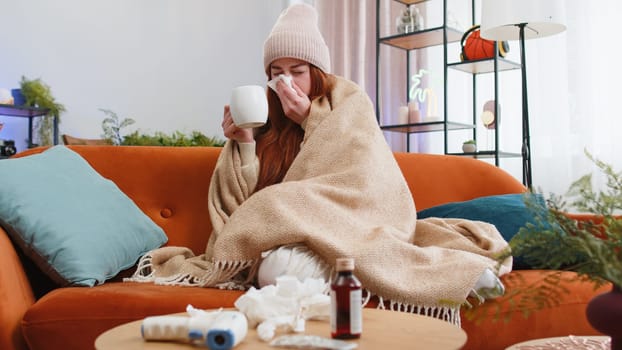 Young ill sick woman suffering from cold or allergy drinking hot tea at home apartment. Redhead girl wear hat wrapped in plaid blows sneezes wipes snot into napkin sitting on sofa in room. Lifestyles