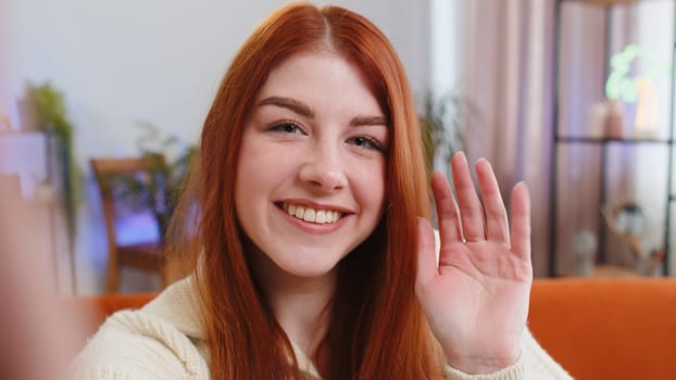 POV of young woman making online selfie video call talking waving hand hello, looking at camera at home apartment indoors. Communication, meeting. Redhead girl blogger making smartphone conversation
