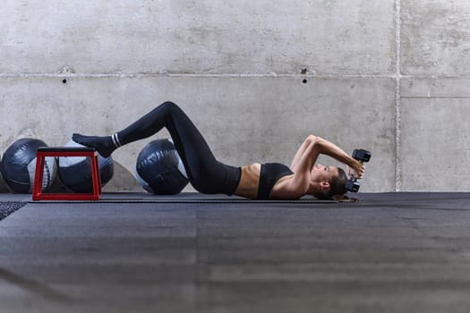 A fit woman is lying on the gym floor, performing arm exercises with dumbbells and showcasing her dedication and strength
