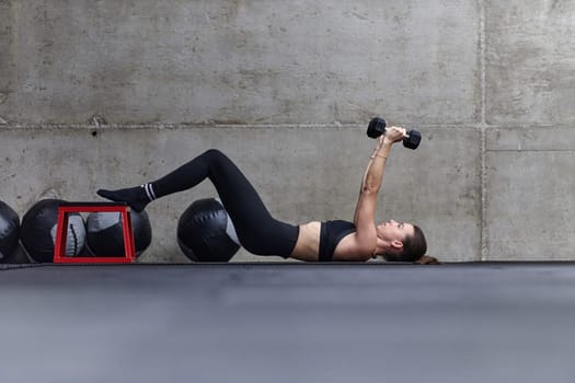 A fit woman is lying on the gym floor, performing arm exercises with dumbbells and showcasing her dedication and strength