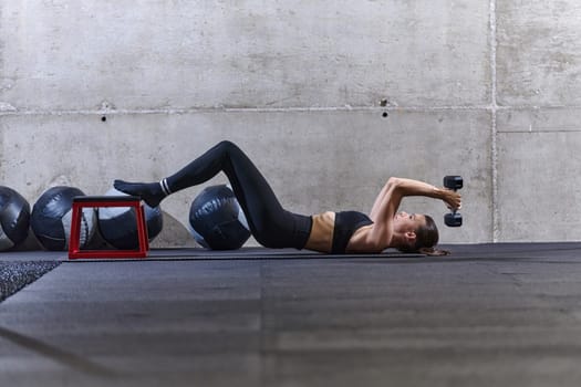 A fit woman is lying on the gym floor, performing arm exercises with dumbbells and showcasing her dedication and strength