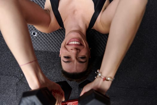 A fit woman is lying on the gym floor, performing arm exercises with dumbbells and showcasing her dedication and strength