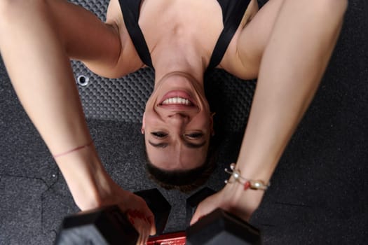 A fit woman is lying on the gym floor, performing arm exercises with dumbbells and showcasing her dedication and strength