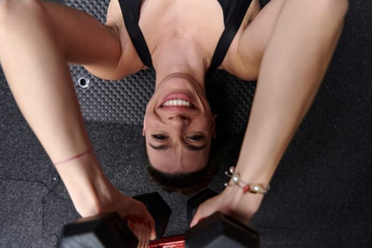 A fit woman is lying on the gym floor, performing arm exercises with dumbbells and showcasing her dedication and strength