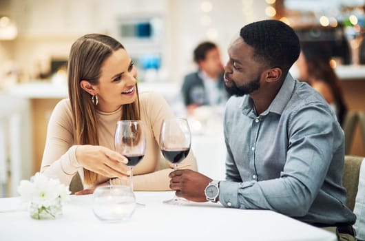 Love, wine and interracial couple on date for fine dining, restaurant or valentines celebration together. Black man and happy woman with romance alcohol glasses, celebrate and luxury in night bokeh.