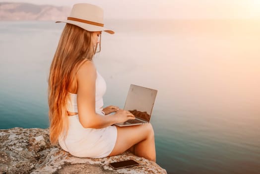 Digital nomad, Business woman working on laptop by the sea. Pretty lady typing on computer by the sea at sunset, makes a business transaction online from a distance. Freelance, remote work on vacation