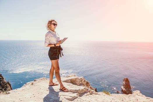 Successful business woman in yellow hat working on laptop by the sea. Pretty lady typing on computer at summer day outdoors. Freelance, travel and holidays concept.