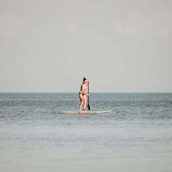 Sea woman sup. Silhouette of happy middle aged woman in rainbow bikini, surfing on SUP board, confident paddling through water surface. Idyllic sunset. Active lifestyle at sea or river