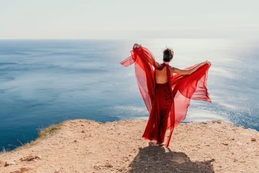 Side view a Young beautiful sensual woman in a red long dress posing on a rock high above the sea during sunrise. Girl on the nature on blue sky background. Fashion photo.