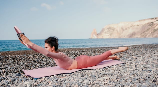 Middle aged well looking woman with black hair doing Pilates with the ring on the yoga mat near the sea on the pebble beach. Female fitness yoga concept. Healthy lifestyle, harmony and meditation.