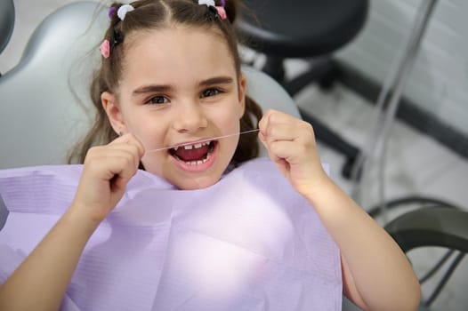 Close-up portrait of beautiful little girl using dental floss, brushing her teeth, sitting in the dentist's chair and smiling cutely, looking at the camera. Oral hygiene concept for caries prevention