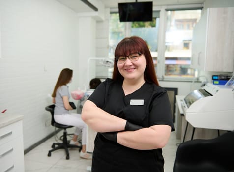 Caucasian positive young woman dentist doctor, dental assistant, orthodontist, maxillofacial surgeon dressed in black medical uniform, smiling at camera, standing with her arms folded in dental clinic