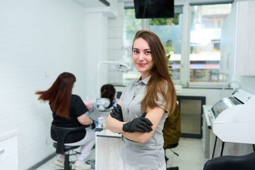 Caucasian charming woman 25-20 years old, wearing gray medical uniform, confident female dentist doctor, orthodontist, maxillofacial surgeon smiles at camera, stands with folded arms in dental clinic