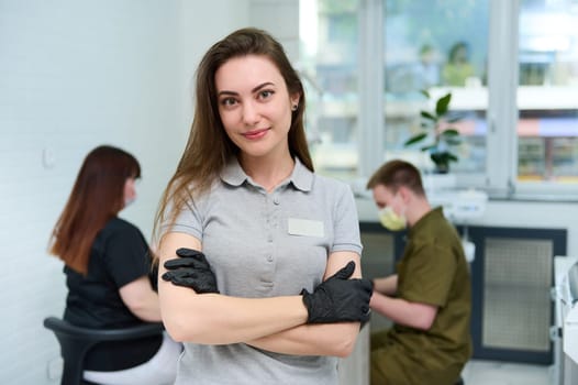 Confident portrait of Caucasian successful committed young female dentist doctor looking at camera, standing in dental office, on the background of dental hygienist and assistant working with patient