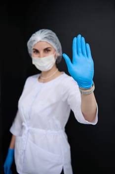 Focus on hand in blue protective latex glove, of blurred woman doctor, surgeon, medical staff, healthcare worker in white lab coat and protective medical mask, shows her hand palm to camera. STOP sign