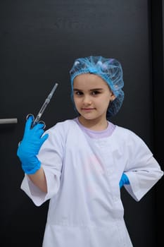 Conceptual portrait child girl playing dentist, dressed like doctor in white lab coat, smiling looking at camera, holding syringe with dental anesthetic, isolated black background. Future profession