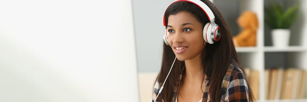 Smiling young black woman in headphones sitting at desk. Woman freelancer working on computer and writing notes in notebook and watching webinar