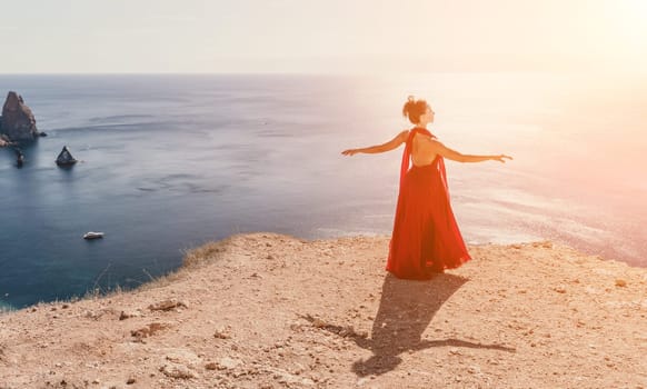 Side view a Young beautiful sensual woman in a red long dress posing on a rock high above the sea during sunrise. Girl on the nature on blue sky background. Fashion photo.