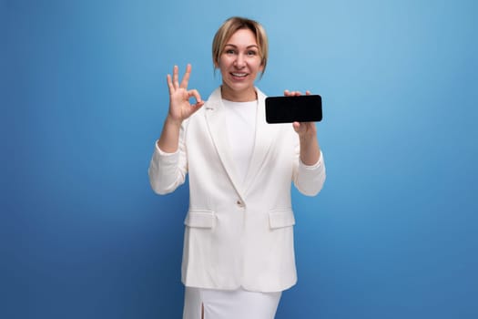 joyful young caucasian blondie lady in elegant jacket holding smartphone with screen mockup.