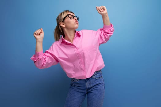 young pretty caucasian blonde secretary woman with ponytail hairstyle, glasses and in a pink shirt on a blue background with copy space.