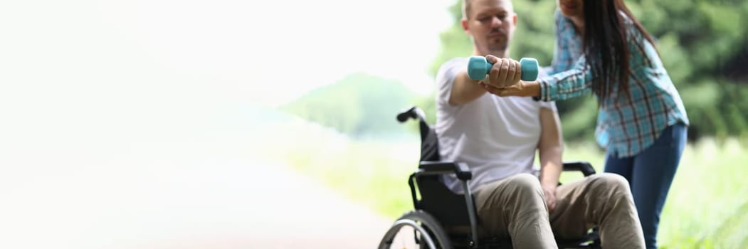 Female physiotherapist helps disabled man in wheelchair to lift weight of arm in park. Rehabilitation and exercises with dumbbells concept