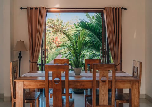 Wooden table with chairs at window with nature view. Potted tropical plant on table, summer balinese villa