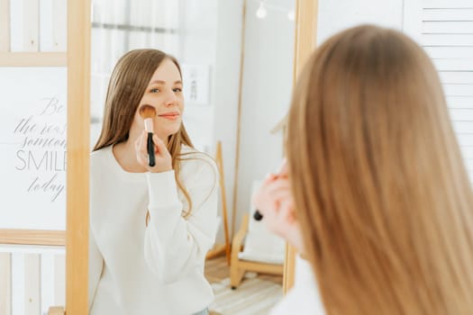 Beautiful smiling young caucasian woman with long hair standing near mirror, doing makeup and looking at reflection in living room at home.