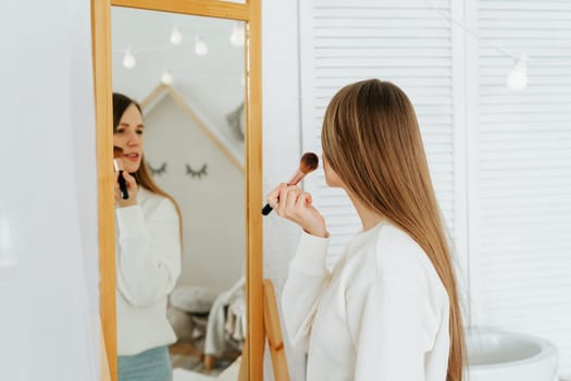 Beautiful smiling young caucasian woman with long hair standing near mirror, doing makeup and looking at reflection in living room at home.
