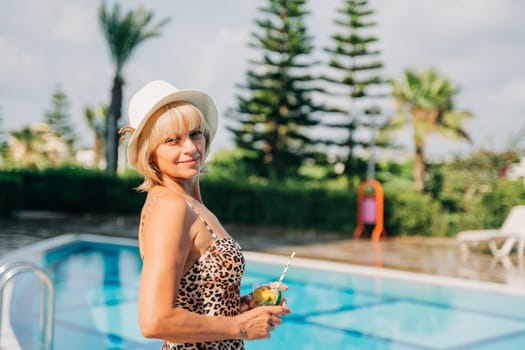Portrait of happy mature adult woman near the pool and looking at camera. Beautiful smiling caucasian senior woman in hat drink lemonade cocktail enjoying summer vacation.