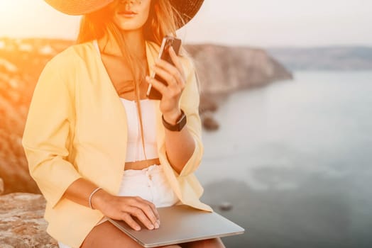 Successful business woman in yellow hat working on laptop by the sea. Pretty lady typing on computer at summer day outdoors. Freelance, travel and holidays concept.