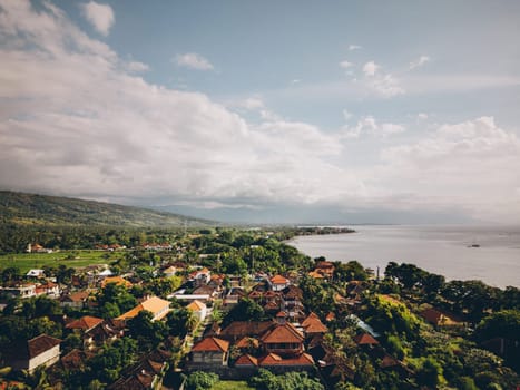 A pretty tropical town sits by the ocean, surrounded by trees and hills. The peaceful scene has a blue sky, fluffy clouds, and shimmering water