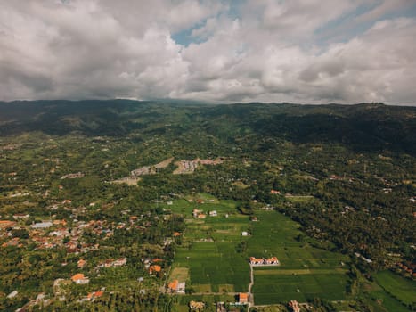 Aerial view of green valley with rolling hills, trees, and houses. Serene atmosphere with blue sky, fluffy clouds, and a winding dirt road. Idyllic glimpse into rural life surrounded by natural beauty