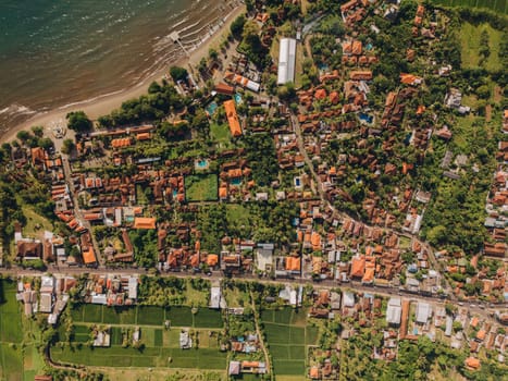 Top aerial view of pretty tropical town sits by the ocean, surrounded by trees and hills. The peaceful scene has a blue sky, fluffy clouds, and shimmering water