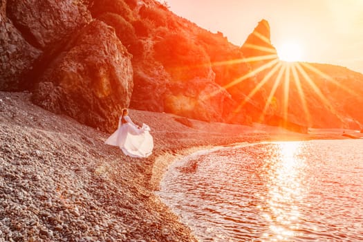 Mysterious woman silhouette long hair walks on the beach ocean water, sea nymph wind listens to the wave. Throws up a long white dress, a divine sunset. Artistic photo from the back without a face.