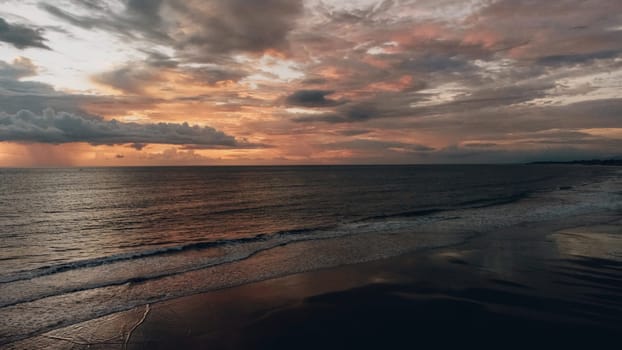 Aerial view of beautiful sunset at the beach. The sky is a mix of grey, black, and orange-red. Waves crash against the shore, reflecting the light