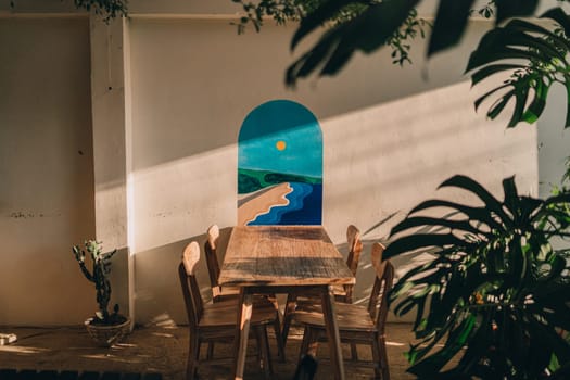 Outdoor summer table in tropical garden. Wooden table with chairs at white wall with decorative painting.