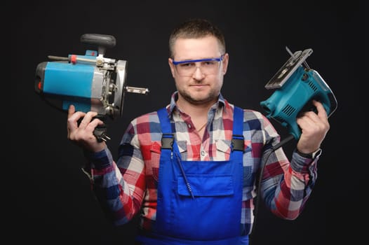 caucasian repairman in uniform and goggles holds electric repair tools. Handyman with tools on black studio background.