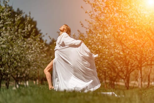 Blond blooming garden. A woman in a white dress walks through a blossoming cherry orchard. Long dress flies to the sides