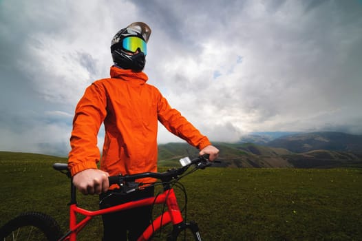 Portrait of a male cyclist in an orange windbreaker and full face helmet with goggles. against the backdrop of a mountainous area and an epic sky.