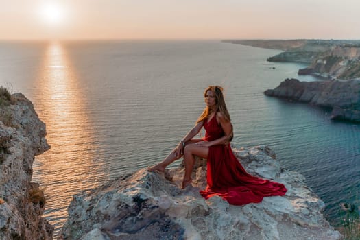 Woman sunset sea red dress, side view a happy beautiful sensual woman in a red long dress posing on a rock high above the sea on sunset